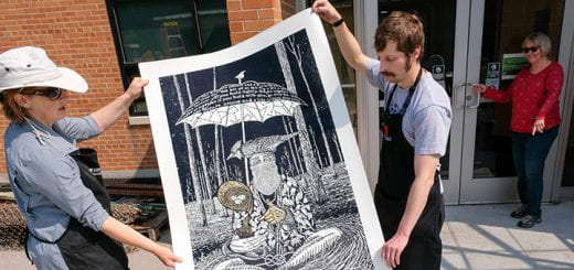 Two artist carry a large relief print that depicts a person sitting on the ground holding a bird underneath an umbrella with text, "We are all in this together...hold with care...nurture nature and a life is..."