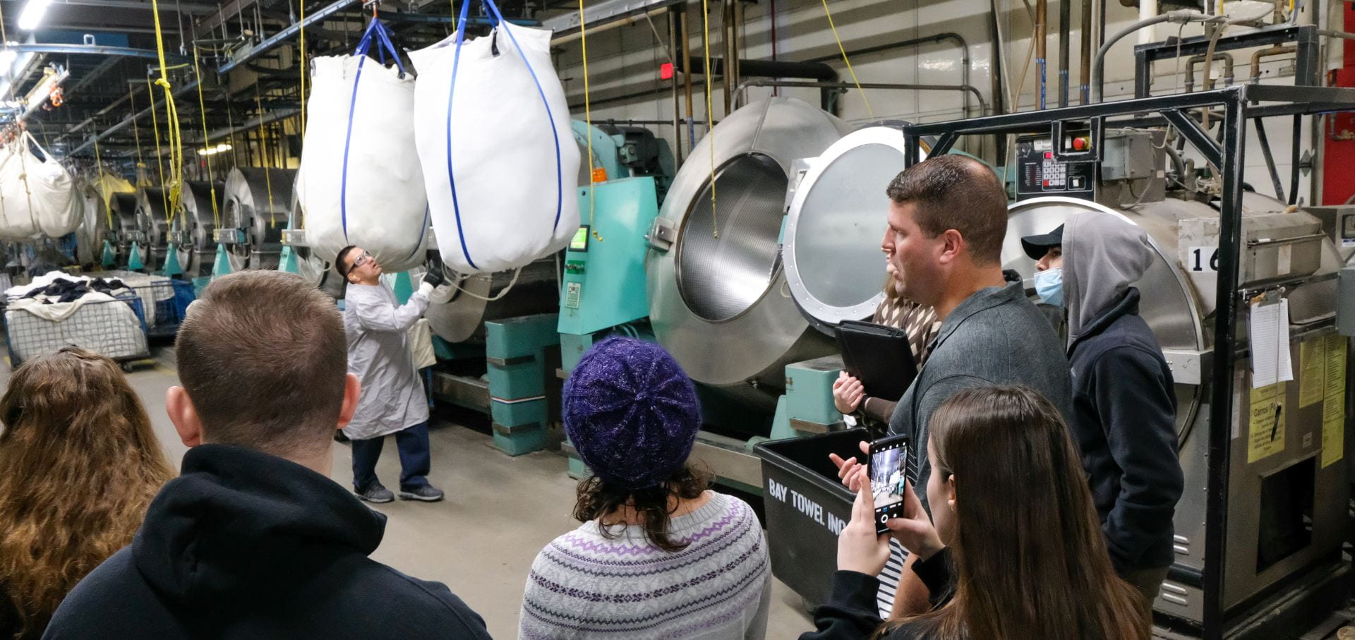 In the foreground, Shane Suda, Vice President of Plant Operations at Bay Towel in Green Bay, explains how commercial laundry is washed to UW-Green Bay business students as an employee moves a large laundry bag using a crane to position the laundry over an extremely large washing machine.