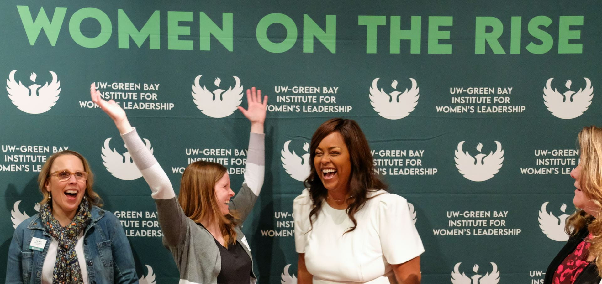 Three females stand in front of the "Women on the Rise" photo backdrop as one female raises her arms up in the air, which makes everyone laugh.