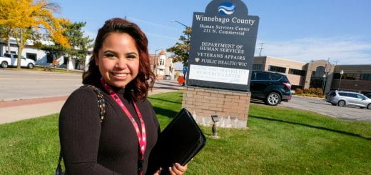 Hannah stands outside on a blue sky, sunny day in front of the sign at Winnebago County Department of Human Services.