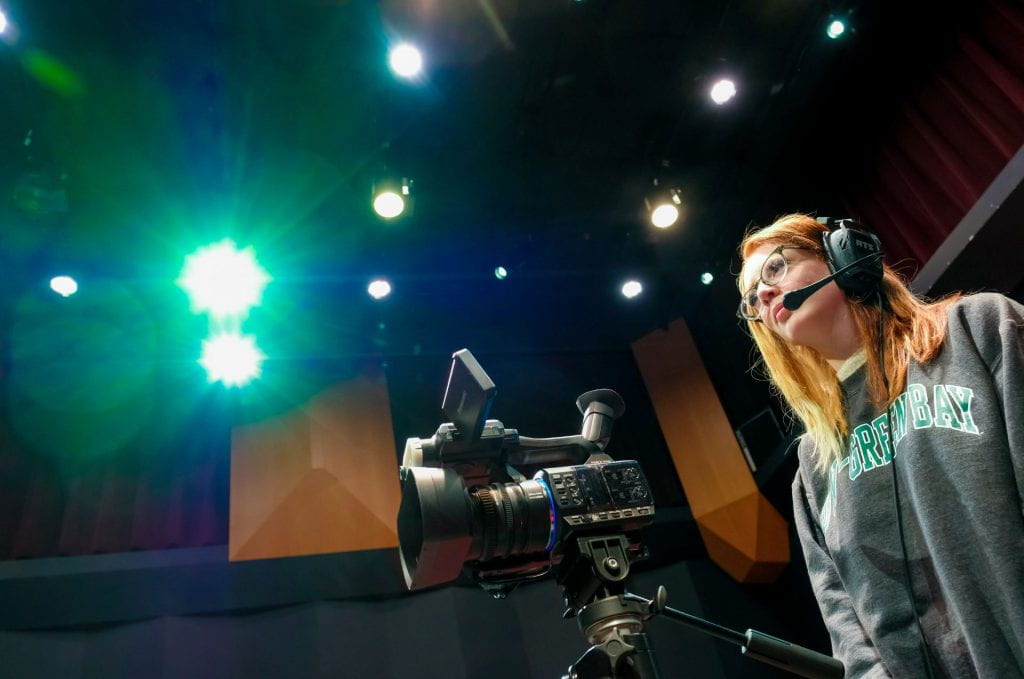 A UW-Green Bay Communication student runs a video camera and behind her are two green and purple rays of light coming of the ceiling studio lights during the filming and production of a live television talk show for their final in Lecturer Mike Schmitt’s Video Production class in the Jean Weidner Theater on April 18, 2022.