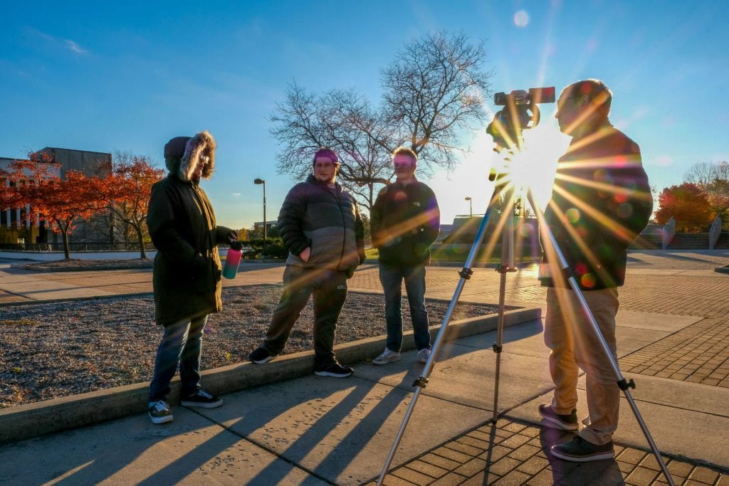 The sun is setting and the rays light up Assistant Professor Justin Kavlie and the video camera on a tripod that he is using to teach camera techniques to students in an outside lab during Advanced Video Production class in peak fall color on Oct. 26, 2022 at the UW-Green Bay, Green Bay campus. 