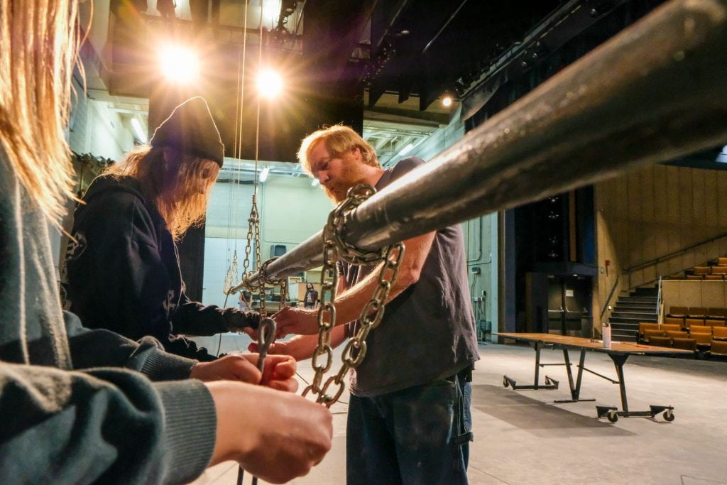 Two bright theatre lights shine rays of light over Instructor David Cook as he shows two theatre students how to safely rig the flying bar to the theatrical rigging system on stage during Stagecraft Class at the University Theatre on Dec. 9, 2022. 