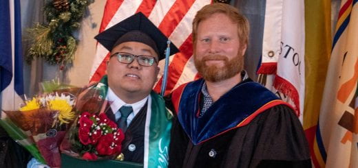 Shue Vang, graduating with a Bachelor of Arts degree in Democracy & Justice Studies, wearing his cap and gown and holding a bouquet of flowers poses in front of the American flag with Jon Shelton, an associate professor and chair of democracy and justice studies at UW-Green Bay.