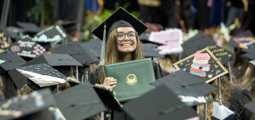 A smiling and standing female graduate holds up her diploma for her family to see that she graduated as she is surrounded by other graduates in a sea of graduation caps during UW-Green Bay's 2023 Spring commencement ceremony.