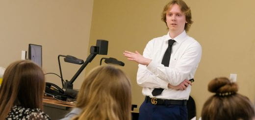 Erik Elliott, a recent graduate of the Management BBA program and currently a banker at BMO Bank, is standing in front of a classroom full of business students, wearing a blue tie and a white shirt as he talks to students about the importance of getting involved on campus during the Student Association of Management meeting.