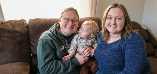An online Organizational Leadership student poses with his one year old son and wife as they smile from the couch in their family room at their home in Pulaski,